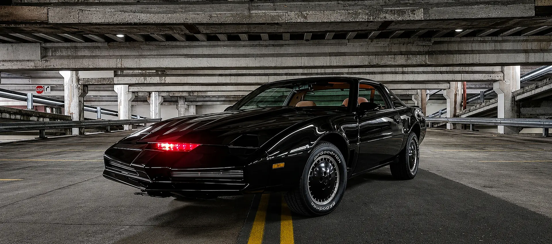 A front three-quarter view of a screen-accurate Knight Rider KITT replica parked inside a concrete parking garage, showcasing its glossy black finish and the active, glowing red scanner light on the nose.
