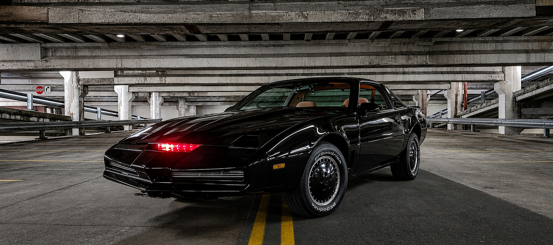 A front three-quarter view of a screen-accurate Knight Rider KITT replica parked inside a concrete parking garage, showcasing its glossy black finish and the active, glowing red scanner light on the nose.